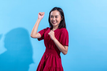 An excited Asian woman in a red dress raises both fists in the air while cheering enthusiastically....