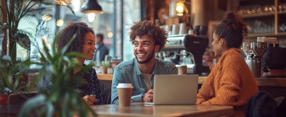 Young man and two women enjoying a friendly conversation in a cozy coffee shop with laptops and coffee cups