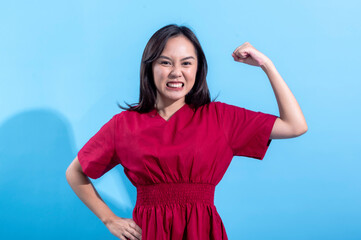 An Asian woman wearing a red dress is flexing her arm, showing strength and confidence, while smiling at the camera. The background is a bright light blue