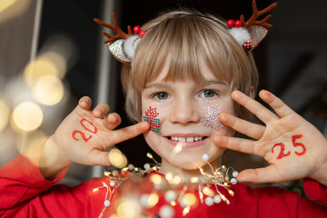 cheerful child holds his palms near face on which numbers of new year 2025 are written. boy is...