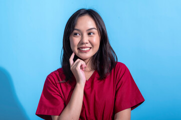 Asian woman in a red dress stands against a light blue background. She is smiling warmly while winking and lightly tapping her cheek with her finger, conveying a playful and confident expression