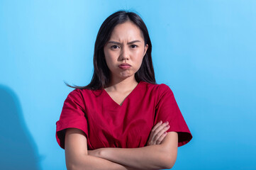Fototapeta premium An Asian woman wearing a red dress stands with her arms crossed and an upset expression, cheeks puffed out, against a light blue background