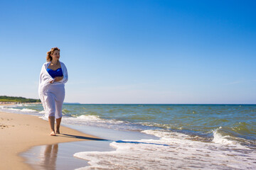 Summer vacations. Beautiful young woman in blue swimsuit covered with white scarf walking on sand by sea on beach on sunny day. 