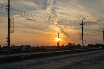 Orange sunset against of a power transmission tower. Cloudy sky. Asphalt road, pillars, horizon in the industrial part of the city. The Far North of Russia.