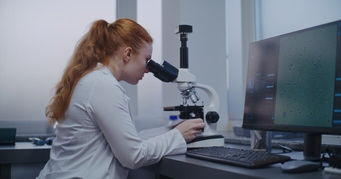 Medical science lab: Female microbiologist looks under microscope. Professional scientist does analysis of sample using high-tech equipment. PC screen shows image of cells or bacteria. Microbiology.