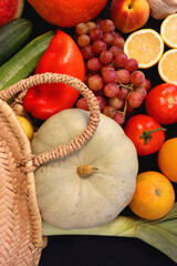 Round straw bag and various seasonal fruits and vegetables on dark background. Summer and fall produce. Top view.