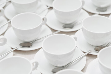 white empty porcelain cups on saucers with spoons in several rows on white tablecloth, serving tables for banquet in restaurant or cafe, pattern, background