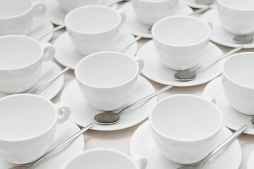 white empty porcelain cups on saucers with spoons in several rows on white tablecloth, serving tables for banquet in restaurant or cafe, pattern, background
