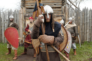 Group of medieval armored warriors standing in front of wooden fort entrance, holding various shields and weapons while wearing helmets and chainmail ensemble - Powered by Adobe