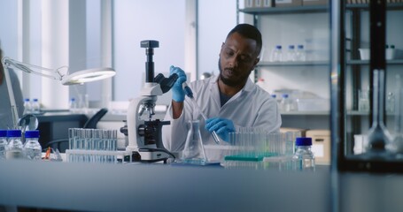 Medical science lab: African American microbiologist takes liquid from test tube, looks under microscope, does analysis of sample using high-tech equipment. Female scientist works in the background.