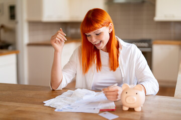 In a bright kitchen, a cheerful woman with bright orange hair smiles as she reviews her bills while...