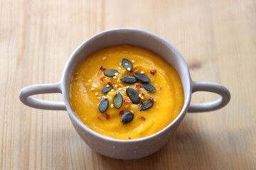 Bowl of creamy pumpkin and potato soup, with pumpkin seeds and chilli flakes toppings. Selective focus, wooden background.