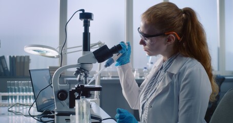Bright medical science laboratory: Female microbiologist looks under microscope, does analysis of test sample, puts on safety goggles. Scientist, biochemist works with high-tech equipment and laptop.