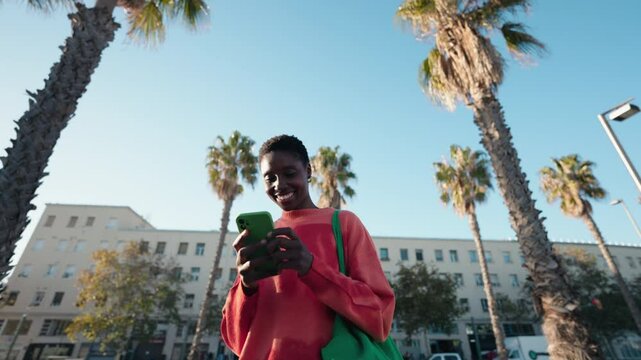 Dark skinned female technology user hold smartphone and laughing while chatting with friends. She walks outdoors on sunny day, surrounded by palms, enjoying light-hearted moment of online communicatio