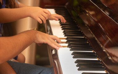 Close-up of an unrecognizable adult and child playing the piano together, focusing on their hands on the keyboard. A warm and intimate moment of musical learning and bonding. 