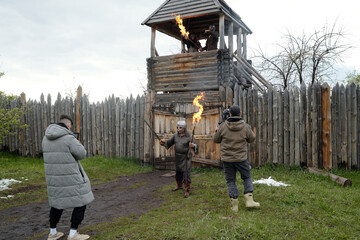 Individuals immersing in medieval reenactment holding flaming torches while photographers capture moment outside wooden fort with tall palisade in background © pressmaster