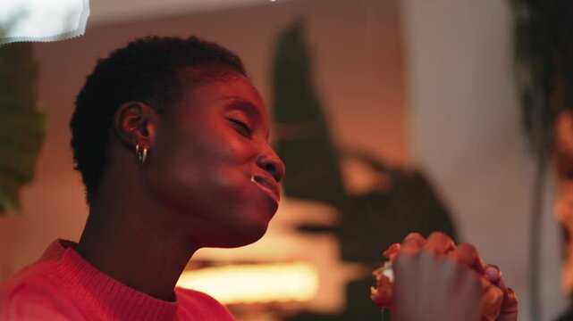 Black woman takes thoughtful bite of loaded burger, savoring each flavor with her eyes closed. Surrounded by warm, ambient glow, she enjoys peaceful moment of indulgence in a cozy, plant-filled space
