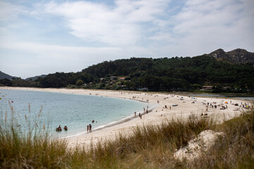 A scenic beach with calm turquoise water, white sand, and people relaxing. Lush green hills and a partly cloudy sky create a peaceful, natural backdrop.