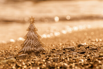 Christmas tree on golden beach sand and blurred glow bokeh on sea waves. Shiny, gold-toned New Year or Christmas background, greeting card, copy space. Celebration winter holidays in warm countries