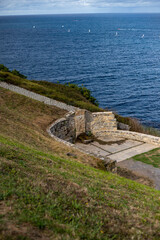 A picturesque coastal scene featuring a stone amphitheater perched on a cliff overlooking a vast ocean. The amphitheater is surrounded by lush green grass and stone walls, providing a stunning panoram