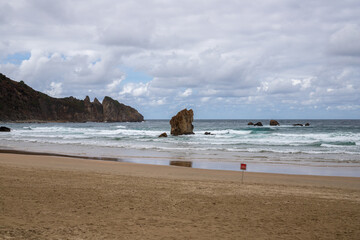 A serene beach scene with crashing waves and a lone rock formation in the distance. The cloudy sky casts a dramatic light on the scene.