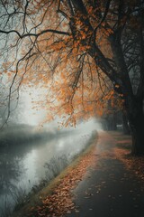 Naklejka premium A tranquil autumn scene with orange leaves lining a quiet pathway by the calm river in the misty morning light.