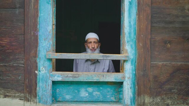 An aged or elderly Muslim Indian Kashmiri man in ethnic attire sitting indoors and looking outside from the frame of the window of an old village wooden house in a mountainous region of Kashmir, India