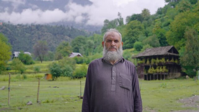An elderly bearded Kashmiri Indian Muslim man or male standing outdoors wearing traditional attire with a green meadow, a scenic mountainous forest region, and a wooden house visible in the background