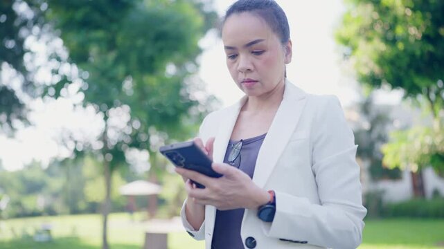 A woman is looking at her cell phone while standing in a park. She is wearing a white jacket and a black shirt