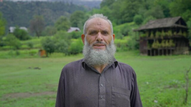 A close up shot of a happy Indian Kashmiri bearded old or aged Muslim man or male in ethnic attire standing outdoors on a green meadow with scenic old village houses facing the camera and smiling