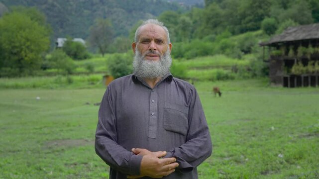 An elderly bearded Kashmiri Indian man or male standing outdoors wearing traditional attire in the green meadow in a mountainous region with scenic village houses, and cattle visible in the background