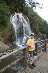 Obraz premium Tourist child, cute kid, fashion boy in Cat Cat village. Landmark of Sa Pa city, northern Vietnam. Vietnamese nature, beautiful waterfall, scenery, landscape. Wooden bridge. Tourism, travel in Sa Pa
