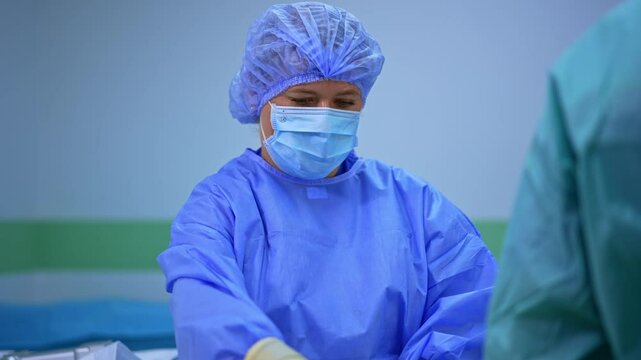 Female scrub nurse wearing blue uniform, cap and mask stands at the surgical stand. Medic prepares sponges and bandages at operation.