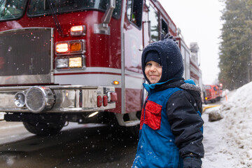 Young boy is smiling near a firetruck while snowflakes fall on a cold winter day. Selective focus