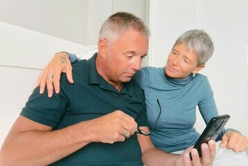 Young couple looking at photos together on smartphone. Loving caucasian lovers holding cell phone, looking at photos or videos together, lying in bed at home, copy space young lovers,smartphone in bed