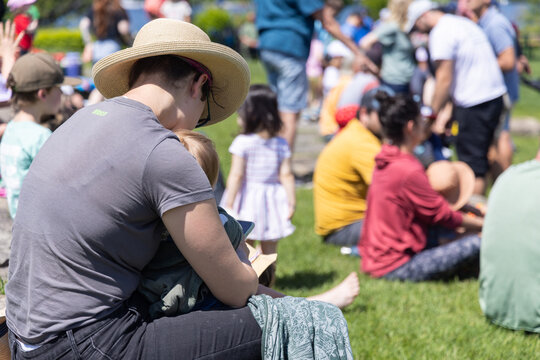 Mother is holding her baby while sitting on a lawn with a crowd of people behind them on a sunny day