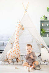 a baby boy in a bright children's playroom with a wigwam and a giraffe playing with wooden toys with cubes, child development © Any Grant