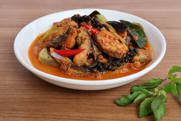 Spicy giant catfish stir-fried with chilli, fresh pepper, kaffir lime leaves, and brinjal eggplant in a cream-colored dish on a wooden table with basil leaves.

