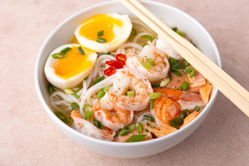 Asian dish with broth, noodles, tiger shrimp, fish, egg and green onions. Traditional Asian soup in a white deep plate. Selective focus, close-up.