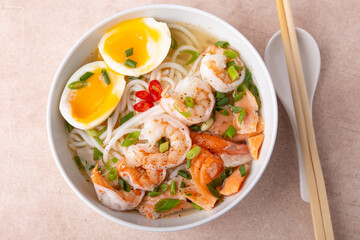 Asian dish with broth, noodles, tiger shrimp, fish, egg and green onions. Traditional Asian soup in a white deep plate. Selective focus, close-up.