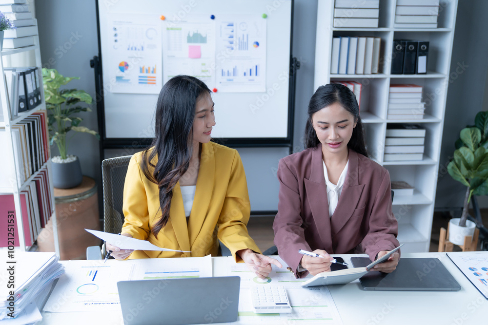 Wall mural two asian businesswomen collaborating in a modern office, analyzing financial documents on a laptop.