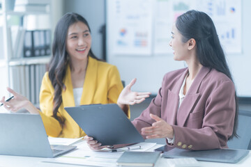 Two businesswomen in a modern office, smiling and having a productive meeting over a document, focused on laptops, working together on a corporate project