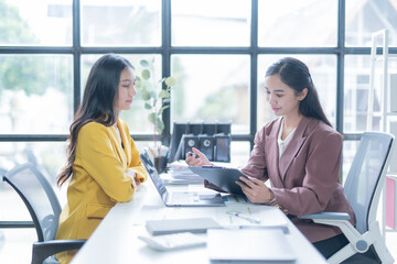 Two young businesswomen are having a meeting in a luminous office, analyzing a financial report with a laptop and a clipboard