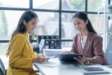 Two asian businesswomen are sitting at a desk in an office, smiling and working together on documents. One businesswoman is holding a clipboard