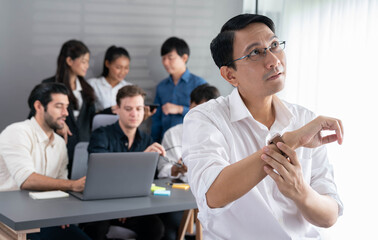 Fototapeta premium Asian businessman portrait check time with his watch with background of his colleague and business team working in office. Office worker teamwork and positive workplace concept. Prudent