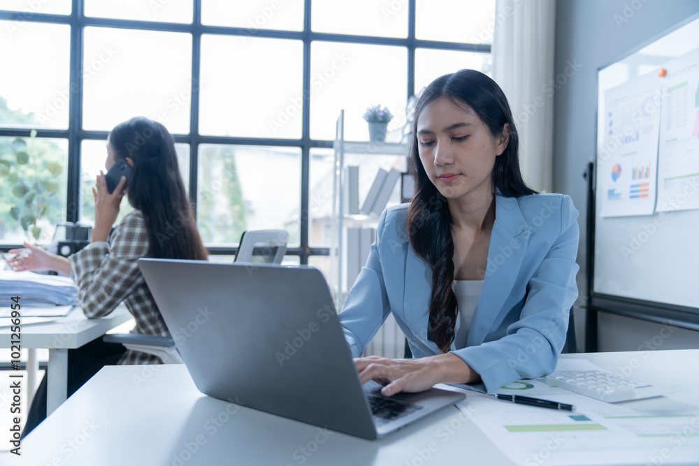 Wall mural young businesswoman wearing a blue blazer is working on her laptop in a modern office with large win