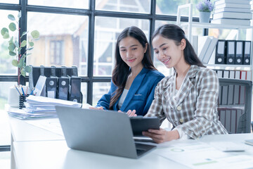 Two young businesswomen collaborate in a modern office, looking at a laptop with charts and documents. Natural light illuminates them as they smile confidently