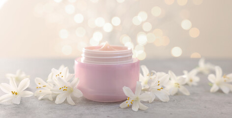 Cream in jar and beautiful jasmine flowers on grey table against beige background with blurred lights, closeup