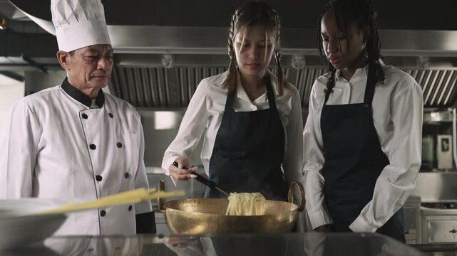 Professional Asian senior male chef in uniform with chef hat teaching cooking methods to female multiracial students listening intently and are ready to learn practical actions in restaurant kitchen. 