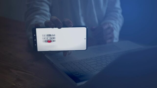 A businessman working on a laptop shows on the smartphone screen a symbolic sign of unification and achieving a goal. The cube is made up of small cubes. Cg